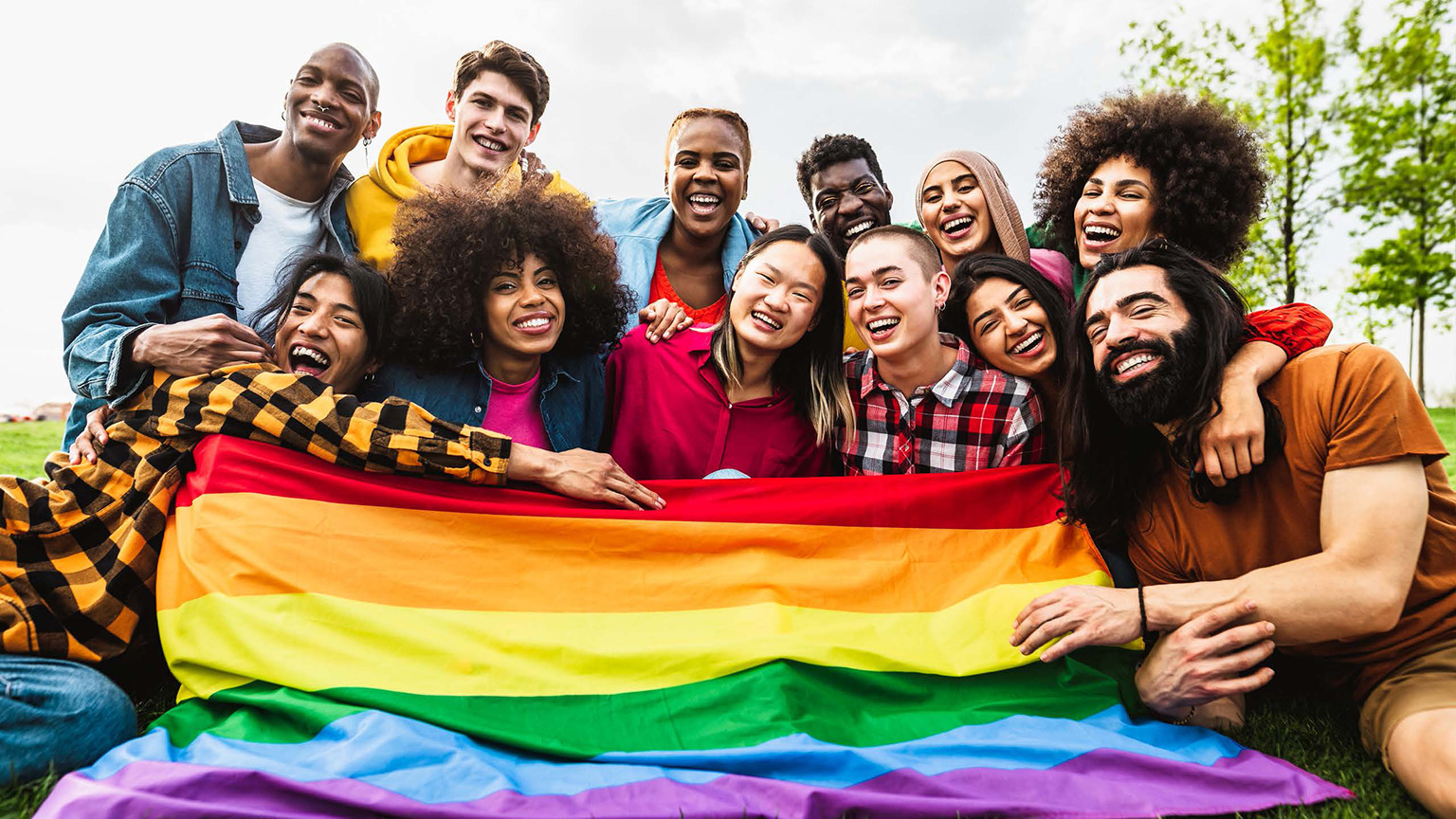 A diverse group of adults joyfully hold a Pride flag in front of them while outside.