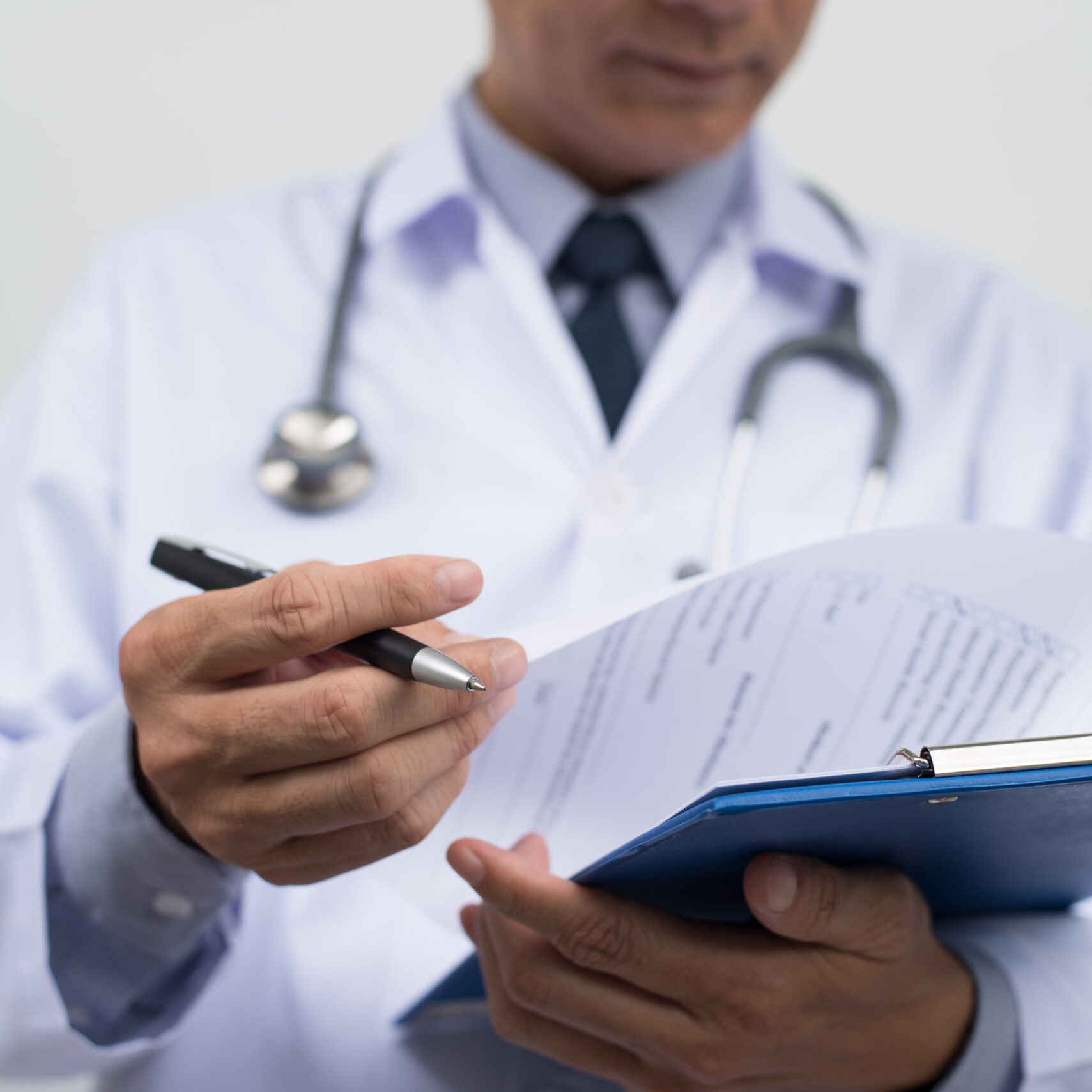 Male doctor on duty in white coat reading patient's information with pen in hand, filling prescription or checklist document, close up, selective focus on pen, health and medical concept.